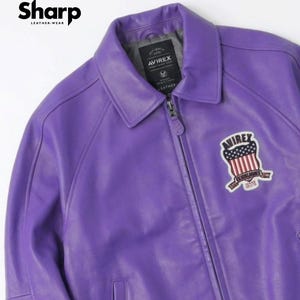 May include: A vibrant purple leather jacket with a collared neckline and a zipper closure. The jacket features a patch with a patriotic design and the brand name "AVIREX". The upper left corner displays the text "Sharp Leather Wear".