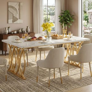 May include: A marble-topped dining table with gold-colored, arched base, set with six cream-colored chairs. The table is set with plates, glasses, and a centerpiece of yellow flowers. The room has a neutral color palette with a patterned rug.