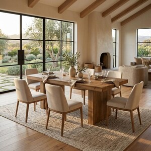 May include: A rectangular wooden dining table with a thick pedestal base, surrounded by six upholstered chairs in a light beige fabric. The table is set with white plates, linen napkins, and two ceramic vases containing green leafy branches.