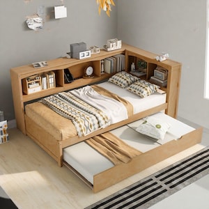 May include: A light-colored wooden daybed with a pull-out trundle bed. The daybed has a built-in headboard with shelves and storage compartments. Pillows and bedding with geometric patterns are visible on the bed.