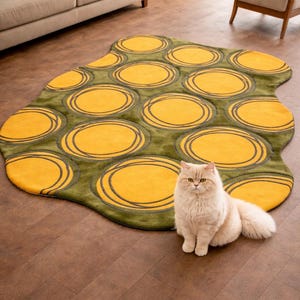 May include: A unique, irregularly shaped rug with a green background and large yellow circles outlined in dark gray. A fluffy, light-colored cat sits in front of the rug on a wood floor. The rug adds a pop of color to the room.