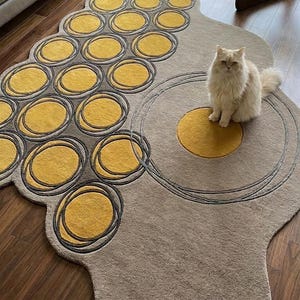 May include: A beige, abstract-shaped rug with a pattern of yellow circles outlined in gray. A fluffy, light-colored cat sits on the rug. The rug is on a hardwood floor, and a sofa is visible in the background.