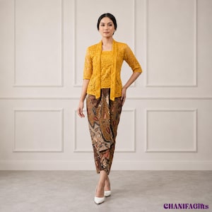 Op de afbeelding: Een mosterdgele kanten kebaya-top met driekwartmouwen gecombineerd met een lange batik sarongrok met een bruin, zwart en crèmekleurig bloemenpatroon. De outfit wordt gedragen met witte puntige hakken. De tekst CHANIFAGifts is zichtbaar.