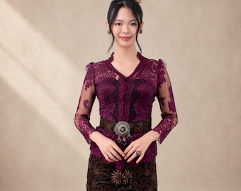 May include: A long-sleeved deep purple lace kebaya top with a V-neckline and front button closure, paired with a dark brown batik sarong featuring floral patterns. A wide, dark, ornate belt with a large silver-toned circular buckle is worn at the waist.