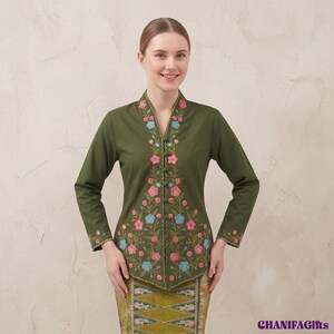 Puede incluir: Una blusa de color verde oliva de manga larga con escote en V, una hilera de botones forrados de tela y un intrincado bordado floral en tonos rosa y azul claro. La prenda incluye puños bordados a juego y una falda estampada.