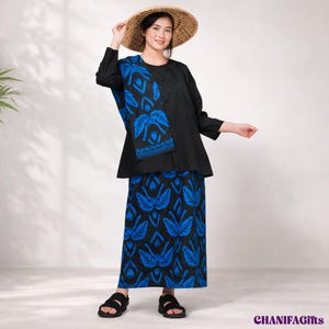 Op de afbeelding: Een set bestaande uit een zwarte tuniek met lange mouwen en een bijpassende rok met een levendig blauw botanisch patroon. De tuniek heeft aan één kant een effen zwart paneel en aan de andere kant een blauw paneel met een patroon.
