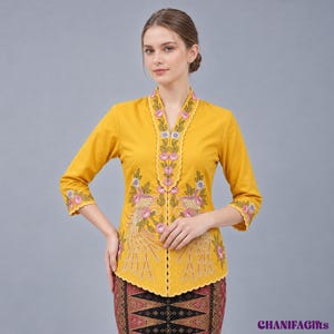 Puede incluir: Una blusa de manga larga color amarillo mostaza con cuello en V, bordes festoneados y bordados florales detallados en rosa, blanco y verde. La prenda presenta un patrón inspirado en un pavo real y se combina con una falda estampada.