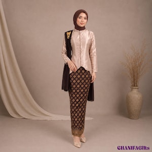 Pode incluir: Uma blusa cor de champanhe de manga comprida com uma fila de botões na frente, combinada com uma saia castanho-escura com padrão batik e detalhes dourados. Um xaile castanho-escuro está drapeado sobre um ombro, preso com um broche dourado.