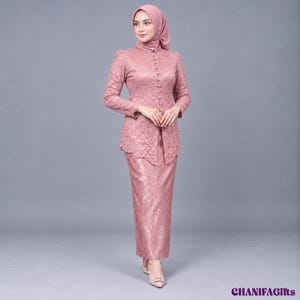 Op de afbeelding: Een oudroze kanten kebaya-set bestaande uit een top met lange mouwen en een hoge kraag, een rij kleine knoopjes aan de voorzijde en een bijpassende rok tot op de grond. De outfit is voorzien van een bijpassende hoofddoek. Tekst: CHANIFAGifts.