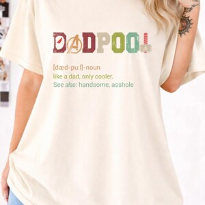 May include: Cream-colored t-shirt with the word "DADPOOL" in colorful block letters. The design includes a definition and the phrase "like a dad, only cooler." The shirt is a casual wear item.