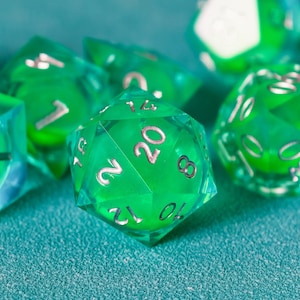 Puede incluir: Primer plano de varios dados poliédricos verdes translúcidos con números color cobre. Los dados están dispersos sobre una superficie turquesa. La atención se centra en un dado de veinte caras, con el número 20 prominentemente mostrado.