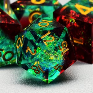 Op de afbeelding: Close-up van een set polyhedrale dobbelstenen. De D20 dobbelsteen is smaragdgroen met gouden cijfers. De binnenkant heeft een glinsterend, iriserend effect. Andere dobbelstenen in de set zijn zichtbaar, sommige met rode en gouden accenten. De dobbelstenen zijn voor tabletop games.