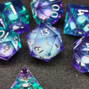 Peut inclure: Un ensemble de dés polyédriques bleus, violets et turquoise. Les dés sont transparents avec un effet tourbillonnant rappelant une galaxie et des chiffres argentés. Différentes formes sont visibles, dont un d20, d6 et d4, parfaits pour les jeux de rôle.