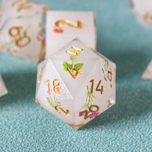 Puede incluir: Un dado transparente de múltiples caras con números dorados y diseños florales. El dado presenta flores incrustadas en tonos de rosa, amarillo y verde. Los números están en una fuente metálica dorada. Otros dados son visibles en el fondo.