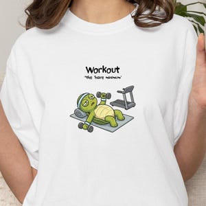 May include: White t-shirt with a cartoon turtle exercising. The turtle is on a gray mat, lifting dumbbells, with a treadmill in the background. Text above the turtle reads "Workout" and "the 'bare minimum'".