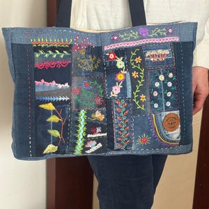 Könnte beinhalten: Jeans-Tote-Tasche mit Patchwork-Design. Die Tasche hat verschiedene gestickte Blumen- und Blättermuster in leuchtenden Farben. Die Tasche hat einen dunkelblauen Riemen. Die Tasche wird von einer Person gehalten, die einen weißen Pullover und eine dunkelblaue Jeans trägt.