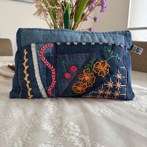 Puede incluir: Un bolso de mano de mezclilla con diseños florales y de cerezas bordados. El diseño de patchwork presenta costuras naranjas, rosas y amarillas. Tachuelas plateadas adornan el borde superior y un pequeño lazo de mezclilla está en el lateral.