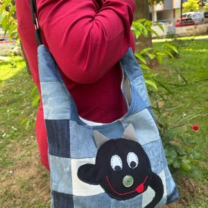 Puede incluir: Un bolso tote de mezclilla patchwork con una cara de personaje de dibujos animados negro. El personaje tiene ojos blancos, cuernos grises y una sonrisa roja cosida con una lengua. El bolso se lleva al hombro.