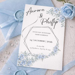 Può includere: Un invito di nozze con i nomi "Aurora & Phillip" in elegante scrittura. L'invito presenta un design floreale con rose azzurre e un bordo geometrico. È presente anche una busta blu abbinata con un sigillo di cera e una rosa blu.
