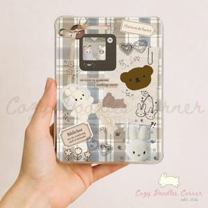 May include: Clear phone case with a plaid pattern and decorative stickers. The stickers include a brown bear, white bunnies, hearts, and the text "we exist in moments, nothing more" and "Biblichor". A small bow is attached to the top.