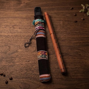 May include: A wooden flute and a black case with colourful woven bands rest on a dark wood surface. The flute is a light brown colour with several finger holes. The case is cylindrical and has a black leather top.