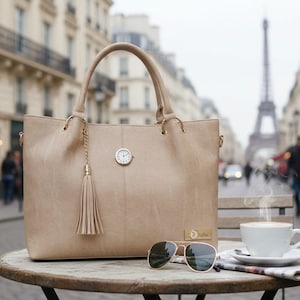 May include: A beige tote bag with a tassel and a small clock charm sits on a table with sunglasses and a cup of coffee. The Eiffel Tower is visible in the background, suggesting a Parisian setting.