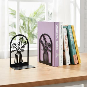 May include: Black metal bookends with a floral design. The bookends feature a vase with flowers and leaves. Several books with colorful spines are placed between the bookends on a wooden surface.