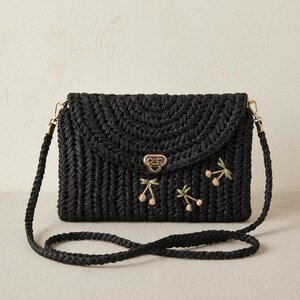 May include: Black woven crossbody bag with a flap closure and gold-tone hardware. The bag features embroidered cherry accents and a braided strap. The bag is approximately 10 inches wide and 7 inches tall.