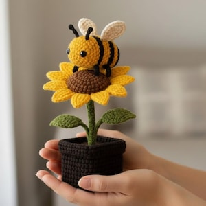 May include: A crocheted bee sits on a yellow sunflower in a black square pot. The bee is yellow and black, with small black antennae and cream wings. The sunflower has a brown centre and green leaves. The pot is held in hands.