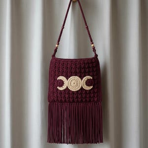 Puede incluir: Bolso de ganchillo burdeos con flecos en la parte inferior y una correa larga. El bolso presenta un diseño de triple luna de color crema en la parte delantera. La correa tiene cuentas de madera. El bolso está colgado de un gancho de madera.