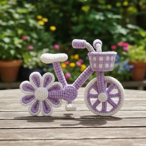 Puede incluir: Una bicicleta de ganchillo hecha a mano en tonos lavanda y blanco. La rueda delantera es una flor grande, y la bicicleta tiene una cesta y un asiento. Se exhibe sobre una superficie de madera con un fondo borroso de vegetación y flores.