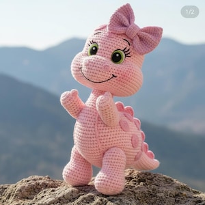 May include: A handmade pink crochet dinosaur toy with a large bow on its head. The dinosaur has a smiling face, green eyes, and pink spots along its back. It is standing on a rock with a blurred mountain backdrop.