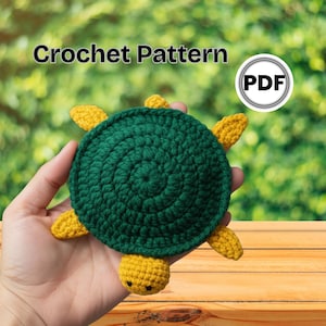 May include: A handmade crochet turtle in shades of green and yellow. The turtle is held in a hand, with the text "Crochet Pattern" and a PDF icon in the background. The turtle has a round green shell and yellow head, feet, and tail.