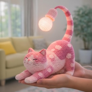 May include: A pink and white crocheted cat lamp with a light bulb at the end of its arched tail. The cat has white polka dots and a smiling face. The lamp is held in a person's hands.