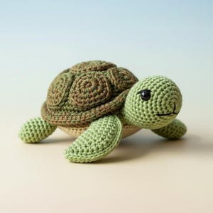 Op de afbeelding: Een handgemaakte gehaakte schildpad in groen- en bruintinten. De schildpad heeft een gedetailleerd schild, een groene kop en vier vinnen. De ogen zijn zwart en de mond lacht. Het is een zacht speelgoed.