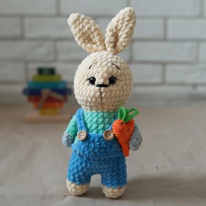 May include: A handmade crochet bunny doll with a cream-coloured head and long ears. The bunny wears blue dungarees with wooden buttons and a turquoise and grey shirt. It holds an orange carrot with green leaves. The background is a white brick wall.