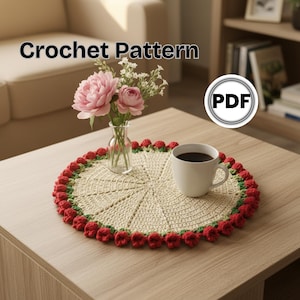 Op de afbeelding: Een gehaakte crème doily met een rode bloemenrand, een kopje koffie en een kleine vaas met roze bloemen op een houten tafel. De doily heeft een cirkelvormig ontwerp. Tekst op de afbeelding luidt "Crochet Pattern" en een "PDF"-pictogram.