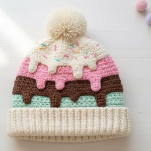 Puede incluir: Un gorro de ganchillo hecho a mano con un diseño de helado. El gorro presenta un pompón color crema y está decorado con capas rosas, marrones y verde menta, que se asemejan a helado derritiéndose. Las chispas de colores añaden un toque lúdico.