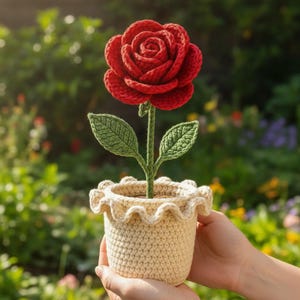 Könnte beinhalten: Eine handgefertigte Häkelrose in einem cremefarbenen Topf. Die leuchtend rote Rose hat grüne Blätter und einen grünen Stiel. Der Topf hat einen gerüschten Rand. Der Hintergrund ist eine verschwommene Gartenszene.
