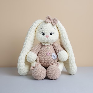 May include: A handmade crochet bunny doll with long, floppy ears and a small bow. The bunny is primarily cream-colored with tan accents on the body, paws, and bow. It has embroidered facial features and a heart-shaped patch.