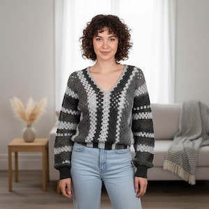 May include: A grey, white, and black crochet sweater with a V-neck. The sweater features a striped pattern on the sleeves and a square pattern on the front. The model is wearing light blue jeans.