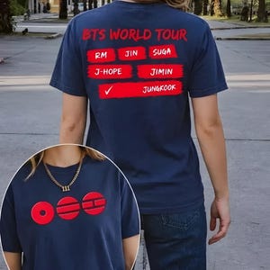 May include: Navy blue t-shirt with red text. The back of the shirt reads "BTS WORLD TOUR" above a list of names. The front of the shirt has the letters "OOO" in red. A gold chain necklace is also visible.