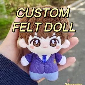 May include: A custom felt doll with brown hair, large eyes, and a purple jacket. The doll has a white shirt and blue trousers. Text on the doll reads "CUSTOM FELT DOLL". The doll is held in a hand.