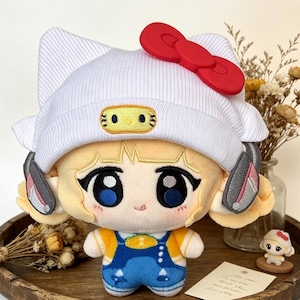 Puede incluir: Muñeca de peluche con un gorro blanco y un lazo rojo. La muñeca tiene grandes ojos azules, pelo rubio y viste una camisa amarilla y un peto azul. La muñeca está sobre una bandeja de madera.