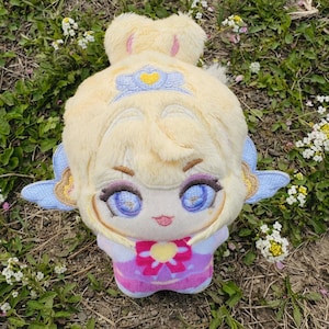 May include: A plush doll with a cartoon-like face, featuring large blue eyes, and a pink and purple outfit. The doll has blonde hair and blue wings. The doll is lying on a bed of grass and small white flowers.