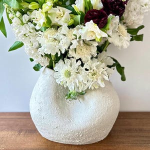 Puede incluir: Un jarrón blanco texturizado lleno de un ramo de flores frescas. Las flores son una mezcla de flores blancas, verdes y borgoña oscuro. El jarrón tiene una forma única y orgánica y está sobre una superficie de madera.