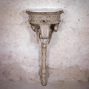 May include: A cream-colored, antique wall-mounted console table. The table features a round top with intricate carvings, supported by a detailed column. The design includes sculpted faces and decorative elements, showcasing a classical aesthetic.