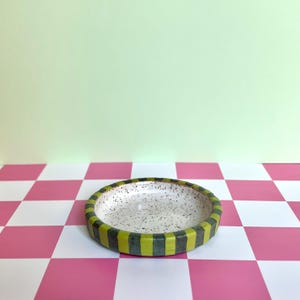 May include: A small, round ceramic dish with a speckled white interior. The exterior rim features a repeating pattern of vertical stripes in alternating shades of lime green and dark olive green, creating a distinct contrast around the edge.