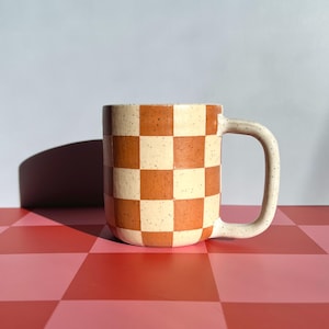May include: A ceramic mug featuring a speckled finish with a repeating checkerboard pattern in alternating shades of terracotta orange and cream. The mug has a cylindrical body and a sturdy, curved handle extending from the right side.