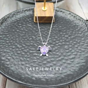 May include: A silver necklace with a turtle pendant. The turtle pendant has a purple and white shell design. The necklace is displayed on a black plate. The text "SAEEJEWELRY" and "Handcrafted Jeweller" are visible.
